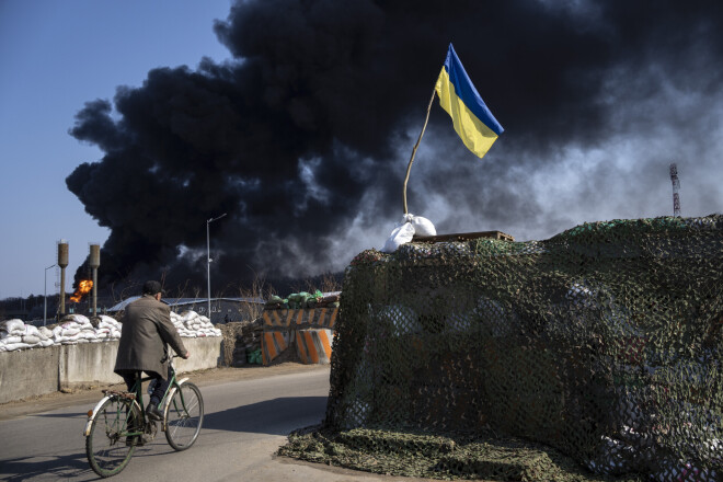 Vīrietis brauc ar velosipēdu, kad pēc Krievijas uzbrukuma no Ukrainas armijas degvielas krātuves paceļas melni dūmi Ukrainas Kijevas pievārtē, piektdien, 2022. gada 25. martā. 