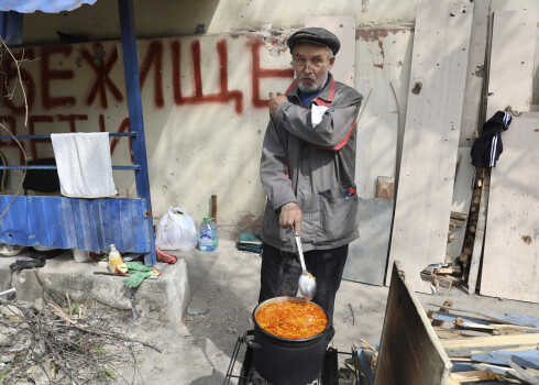 Blakus savai mājai Mariupolē, teritorijā Doņeckas Tautas Republikas valdības pakļautībā, Ukrainas austrumos, trešdien, 2022. gada 4. maijā, ēst gatavo vietējais civiliedzīvotājs pie izkārtnes, kurā lasāms uzraksts “bumbu patversme, bērni.” 