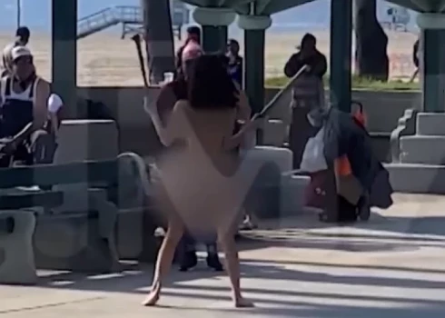 Девушка в "костюме Евы" борется с размахивающей дубинкой дамой; посетители пляжа смотрят с открытыми ртами