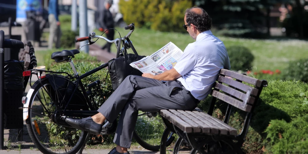 Vīrietis lasa avīzi parkā uz soliņa.