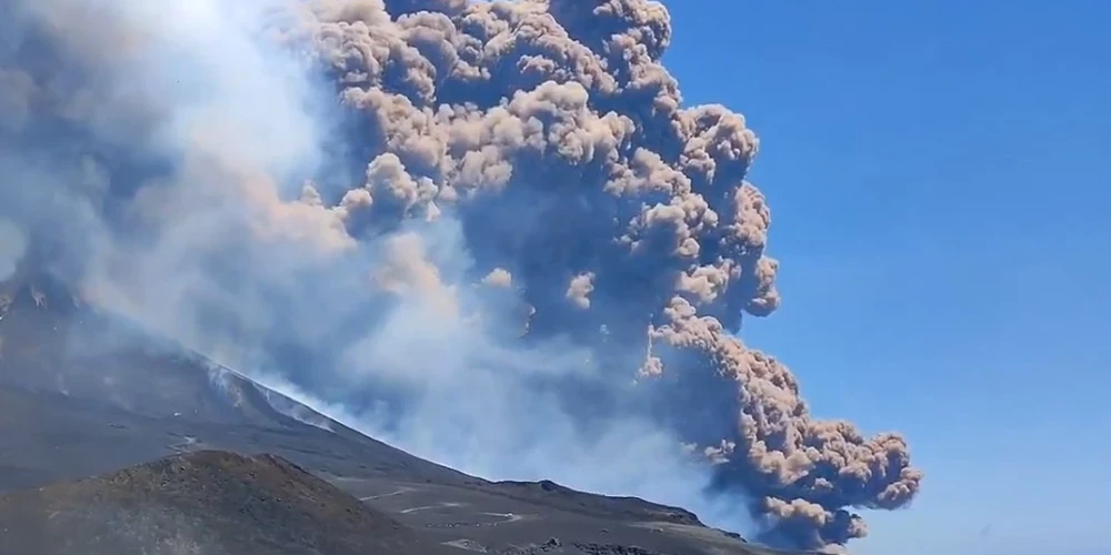 Virs Etna vulkāna redzams milzīgs dūmu un pelnu mākonis, kas paceļas skaidrās, zilās debesīs.