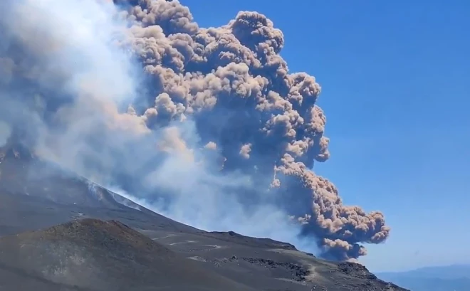 Virs Etna vulkāna redzams milzīgs dūmu un pelnu mākonis, kas paceļas skaidrās, zilās debesīs.