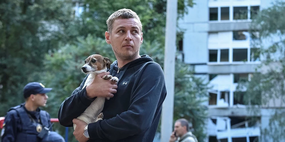 Vīrietis tur rokās savu suni, stāvot pie dzīvojamās ēkas, kas bojāta Krievijas raķešu trieciena laikā Dnipro.