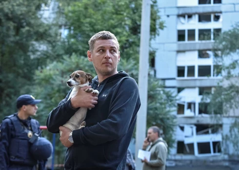 Vīrietis tur rokās savu suni, stāvot pie dzīvojamās ēkas, kas bojāta Krievijas raķešu trieciena laikā Dnipro.