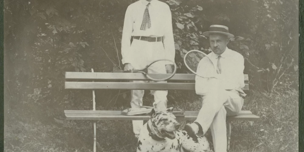 Foto no 20. gadsimta paša sākuma. Latvijā vēl saimnieko Krievijas impērija, Pirmais pasaules karš vēl nav sācies, un kungi var mierīgi spēlēt tenisu. Viņiem pie kājām raibs suns – varētu būt dogs vai vismaz jauktenis ar doga asiņu piejaukumu. Kas ir šie vīri, diemžēl vairs nav zināms, tādēļ varam vien minēt – vācbaltieši vai varbūt veiksmīgi un pie rocības tikuši latvieši, kādu tajā laikā jau bija gana daudz.