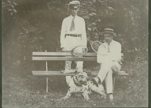 Foto no 20. gadsimta paša sākuma. Latvijā vēl saimnieko Krievijas impērija, Pirmais pasaules karš vēl nav sācies, un kungi var mierīgi spēlēt tenisu. Viņiem pie kājām raibs suns – varētu būt dogs vai vismaz jauktenis ar doga asiņu piejaukumu. Kas ir šie vīri, diemžēl vairs nav zināms, tādēļ varam vien minēt – vācbaltieši vai varbūt veiksmīgi un pie rocības tikuši latvieši, kādu tajā laikā jau bija gana daudz.