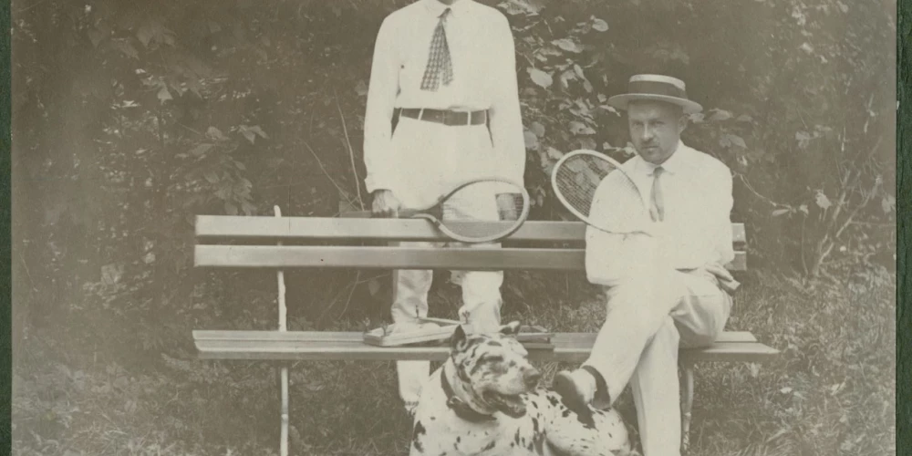 Foto no 20. gadsimta paša sākuma. Latvijā vēl saimnieko Krievijas impērija, Pirmais pasaules karš vēl nav sācies, un kungi var mierīgi spēlēt tenisu. Viņiem pie kājām raibs suns – varētu būt dogs vai vismaz jauktenis ar doga asiņu piejaukumu. Kas ir šie vīri, diemžēl vairs nav zināms, tādēļ varam vien minēt – vācbaltieši vai varbūt veiksmīgi un pie rocības tikuši latvieši, kādu tajā laikā jau bija gana daudz. 