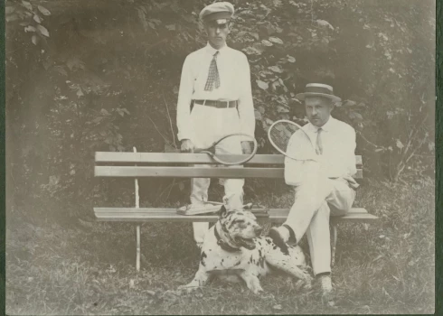 Foto no 20. gadsimta paša sākuma. Latvijā vēl saimnieko Krievijas impērija, Pirmais pasaules karš vēl nav sācies, un kungi var mierīgi spēlēt tenisu. Viņiem pie kājām raibs suns – varētu būt dogs vai vismaz jauktenis ar doga asiņu piejaukumu. Kas ir šie vīri, diemžēl vairs nav zināms, tādēļ varam vien minēt – vācbaltieši vai varbūt veiksmīgi un pie rocības tikuši latvieši, kādu tajā laikā jau bija gana daudz.
