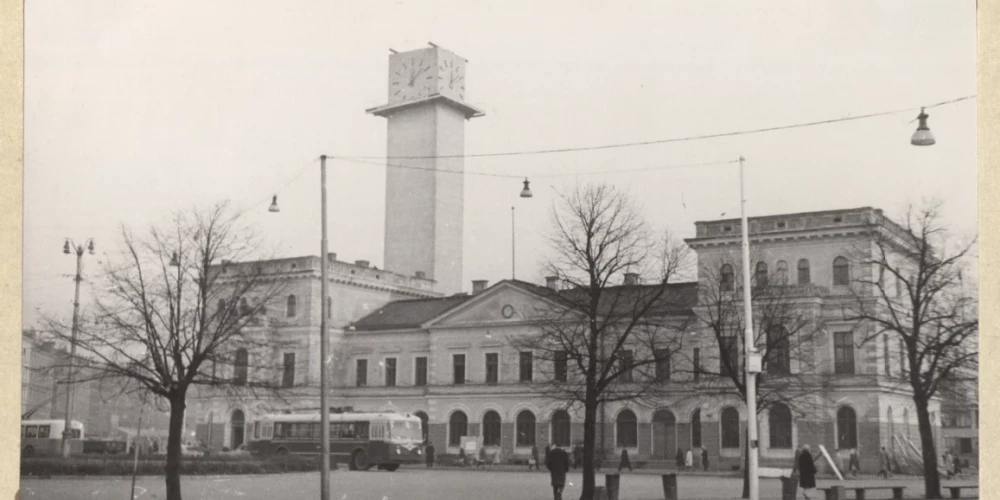 На переднем плане — исторический железнодорожный вокзал Рига (строительство началось в 1858 году; здание вокзала построено по проекту архитектора Иоганна Даниэля Фелско, в 1884–1885 годах вокзал расширили по проекту Генриха Шеля). На заднем плане видна постройка часовой башни нового здания Рижского вокзала (автор фото — Н. Судрабиньш, 1965 год).