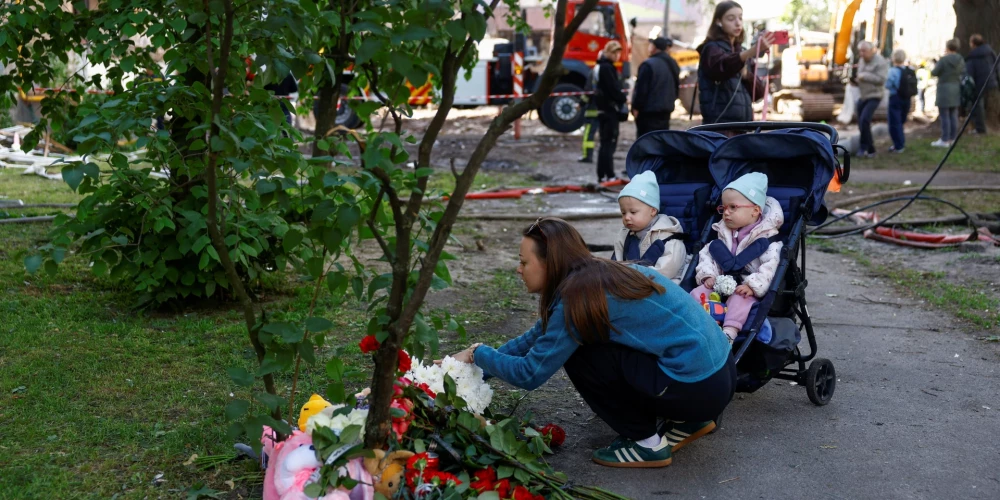 Женщина с детьми возлагает цветы к стихийному мемориалу на месте жилого дома.